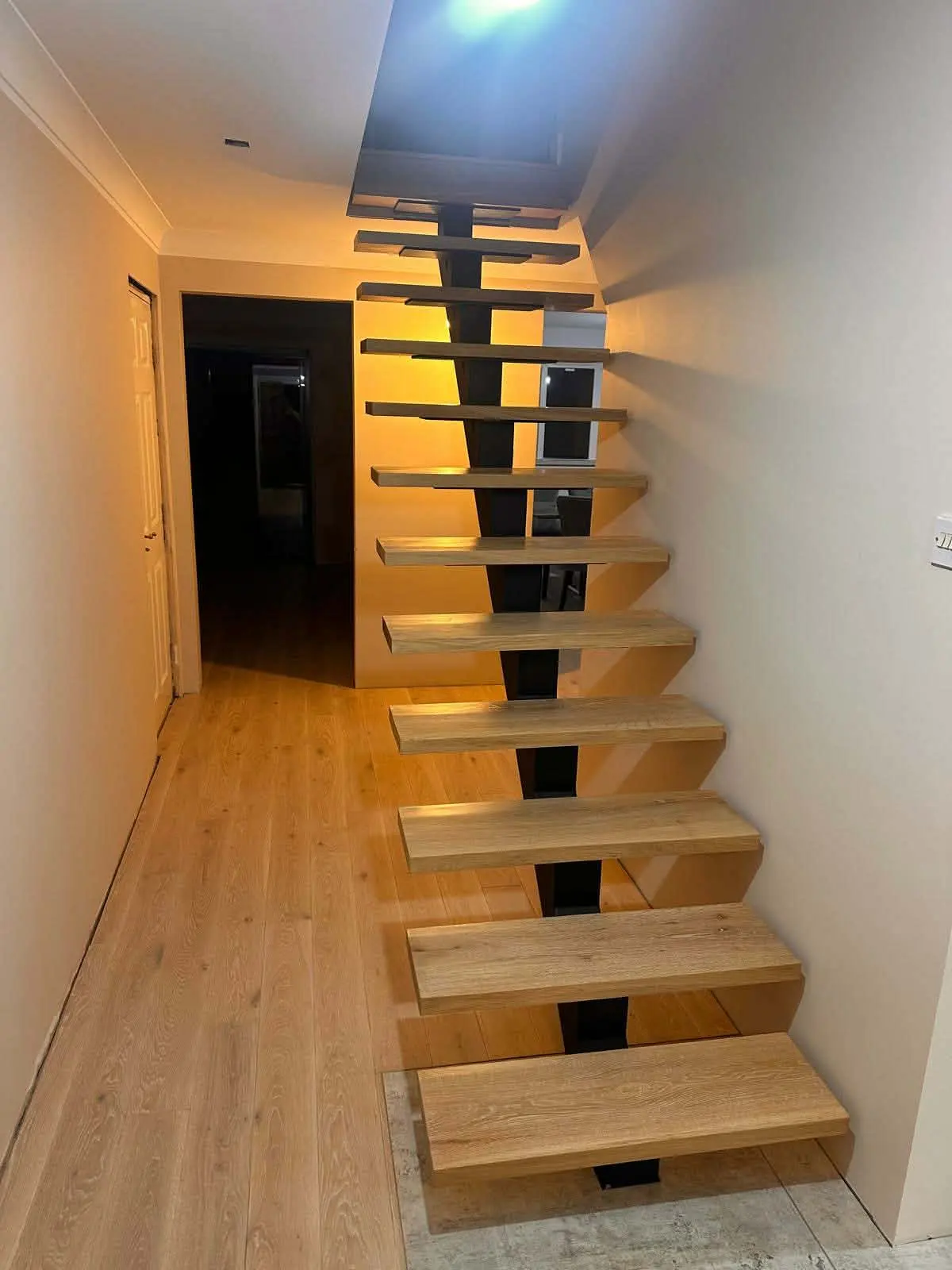 Open-riser staircase with thick oak-toned treads on a dark central spine, lit warmly in a hallway with light flooring